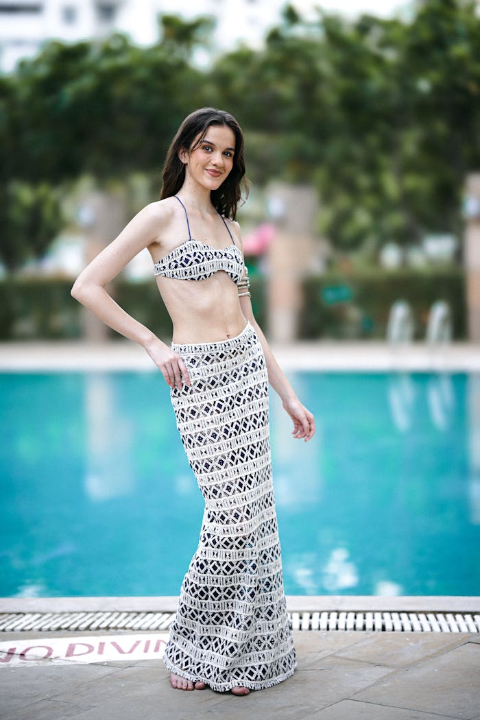Fashionable woman in a crochet dress standing elegantly by a swimming pool.