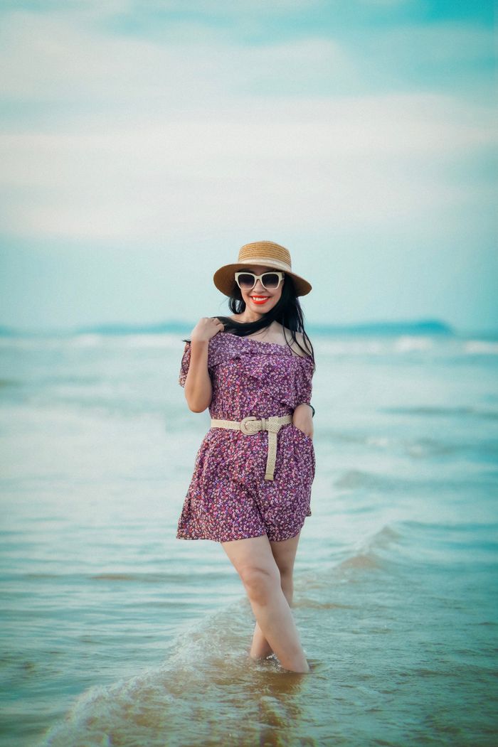 Asian woman in a floral dress with a hat posing by the sea on a sunny day.
